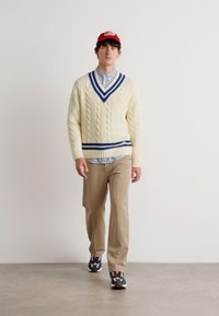 Maglione crema a cavo con accenti blu, indossato sopra una camicia a quadri azzurri, abbinato a pantaloni beige e sneakers con motivo.