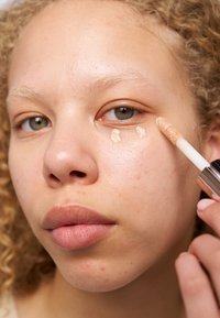 Cream concealer applicator held near the eye, featuring a light beige shade. Skin has a smooth texture with light blemishes visible.