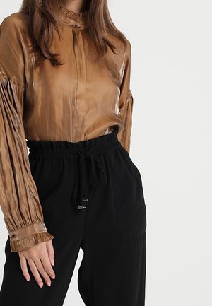 Frau trägt eine glänzende braune Bluse mit langen Ärmeln und Plisseefalten sowie schwarze, hoch taillierte Hose mit Kordelzug.