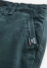 Mörk teal tygbyxor med en slät textur, försedda med knäppning och en "Super Dry"-tagg med röda och vita accenter.