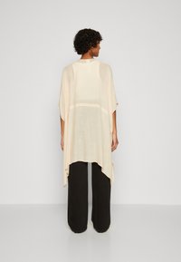 Lys beige kimono-inspirert topp med løse, flytende stoffer, asymmetrisk kant og brede ermer, parret med svarte vidde bukser.