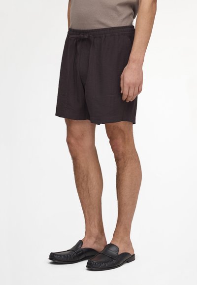 Homme portant un short noir à cordon et des mocassins en cuir noir sans lacets, debout devant un fond blanc uni.
