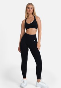 Schwarzer Sport-BH und Leggings aus dehnbarem Stoff mit enganliegendem Design und weißen Logoakzenten. Modell trägt weiße Sneaker.