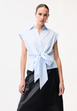 Kvinde iført ærmeløs lyseblå skjorte med store knapper og stor sløjfe foran samt sort lædernederdel, stående mod en hvid baggrund.