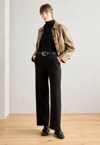 Veste cropped en cuir marron avec un haut noir à col montant, un pantalon large noir et des chaussures noires. Ceinture à boucle dorée, design épuré, détails minimalistes.