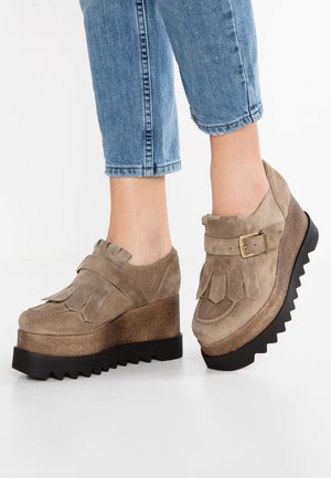 Scarpe in suede beige con una spessa piattaforma in legno frastagliata e dettagli in frange. Presentano una fibbia laterale e una punta arrotondata.