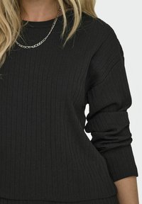 Schwarzer, gerippter Pullover mit langen Ärmeln und Rundhalsausschnitt, aus strukturiertem Stoff mit leichten Raffungselementen an den Bündchen. Silberne Kettenakzent.