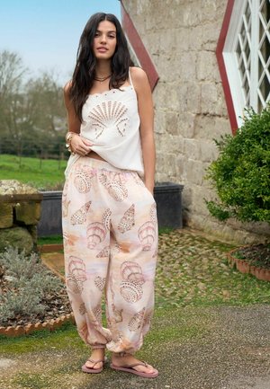 Woman with long dark hair wearing a white sleeveless top with shell cutouts and loose pastel pants with seashell prints, standing outdoors by stone wall.
