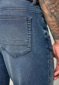 Pantalones vaqueros de mezclilla azul que presentan un bolsillo trasero con un contorno cosido. La tela tiene una textura suave con detalles sutiles de sombreado y desgaste.