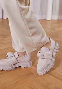 Ljust rosa läderloafers med knutdetalj på remmen, tjock sula och texturerade accenter, i kombination med beige byxor på ett trägolv.