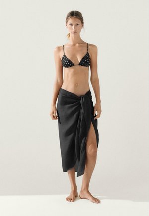 Žena v černém bikinovém topu s puntíky a černém sarongu svázaném v pase, stojící naboso proti jednoduchému světlemu pozadí.