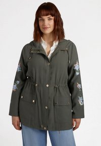 Olivgrön jacka med dragsko i midjan, guldiga detaljer och blommig broderi på ärmarna, med en huva och två framfickor.