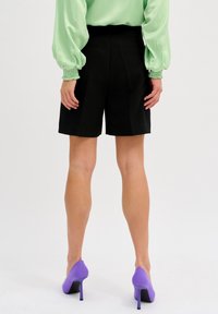 Czarne spodenki o kroju dopasowanym z wysokim stanem i dwoma tylnymi kieszeniami, zestawione z jasną zieloną bluzką i fioletowymi butami na wysokim obcasie.