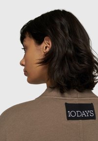 Brauner Strickpullover mit einem rechteckigen schwarzen Etikett auf der Rückseite, auf dem "10DAYS" in weißem Text steht. Weiche Textur und figurbetontes Design.