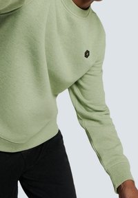 Ljusgrön crewneck sweatshirt med texturerad väv, ribbade ärmslut och en liten hexagonal logotyp på bröstet. Svarta byxor nedanför.