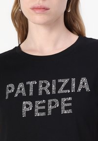 Melna t-kreklas ar tekstu "PATRIZIA PEPE" sudraba strausiem. Īsas piedurknes, apaļa kakla izgriezums, gluda kokvilnas auduma.