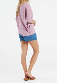 Nainen seisoo paljain jaloin sinisissä farkkushortseissa, väljässä vaaleanpunaisessa kuvioidussa yläosassa ja ruskeissa matalissa sandaaleissa, toinen jalka hieman kohotettuna.