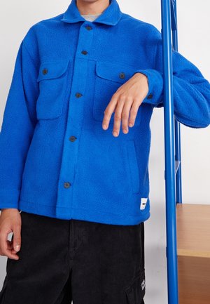 Osoba ubrana w niebieską polarową kurtkę z przednimi kieszeniami, opierająca dłoń na niebieskiej metalowej ramie, ubrana w czarne spodnie.