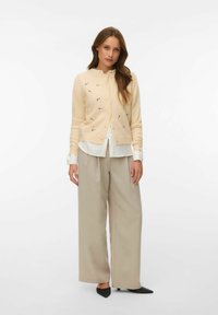 Žena v krémovém květovaném svetru na knoflíky přes bílou košili, béžové kalhoty s širokými nohavicemi a černé špičaté podpatky, stojící na bílém pozadí.