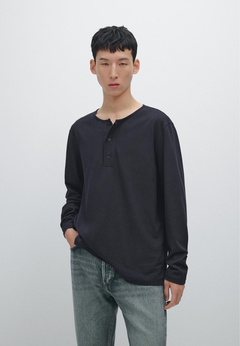 Schwarzes Langarm-Henley-Shirt mit Rundhalsausschnitt, ausgestattet mit drei Knöpfen und lockerer Passform, kombiniert mit hellblauen Jeans. Einfaches Design.