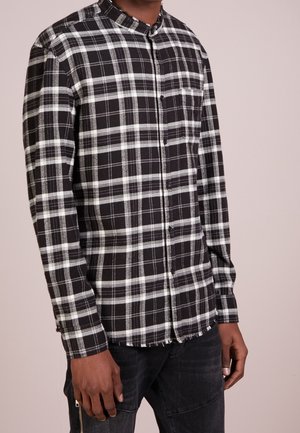 Mężczyzna w czarno-białej, kratowanej koszuli na guziki z kieszenią na piersi, w ciemnych dżinsach na jednolitym tle.