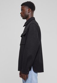 Zwarte wolmix overshirt met een kraag en een frontzak, met lange mouwen en knoopsluiting bij de mouwen. Gemaakt over lichtblauwe jeans.