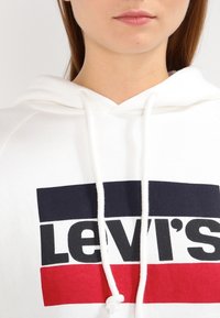 Felpa bianca con cappuccio e cordini, caratterizzata da un grande logo "LEVI'S" in nero, rosso e blu navy sul petto. Tessuto morbido.