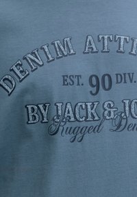 Maglietta di cotone blu con testo stampato in caratteri vari: "DENIM ATTITUDE," "BY JACK & JONES," e "Rugged Denim." Tessuto liscio.
