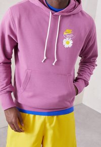 Fialová mikina s bílými šňůrkami, s vyšitým sedmikráskou a logem. V kombinaci se žlutými šortkami a modrým tričkem.