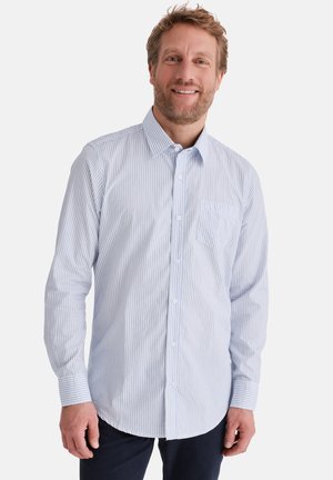 Mann lächelt, trägt ein hellblaues und weißes, dünn gestreiftes Hemd mit langen Ärmeln und dunkle Hosen, vor einem schlichten Hintergrund.