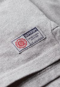 Grijze stof close-up met een genaaid label met "Superdry" in navy en rood, samen met "Vintage Athletic Outfitters." Textuur oppervlakte.
