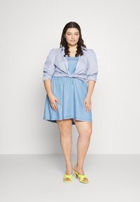 Vestido de ganga azul claro com uma sobreposição de camisa de botão amarrada. Apresenta mangas curtas e uma saia rodada. Combinado com sapatos de salto alto amarelos brilhantes.