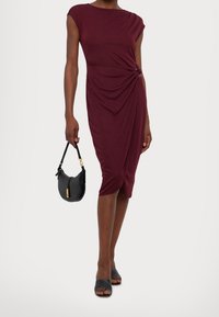 Burgundy, knälång klänning med korta ärmar, draperad fram och med en vriden detalj vid midjan. Accessoarer med en svart handväska och strappy skor.