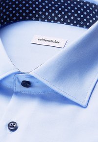 Lichtblauw overhemddas met een puntkraag en eennavyblauwe kleurige binnenkraag met stippen, met zwarte knopen en een zichtbaar merklabel.