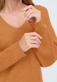 Gehaakte trui in warm oranje, met een V-hals, ribbelmanchetten en een zachte textuur. Close-up van handen die de mouwen aanpassen.