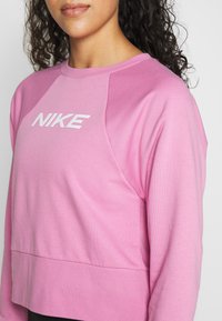 Femme portant un sweat-shirt rose Nike avec un logo blanc, montrant le haut du torse et une partie de cheveux bouclés foncés sur un fond blanc.