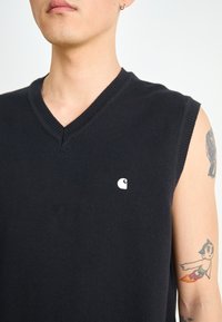 Schwarze ärmellose Strickweste mit V-Ausschnitt. Verfügt über ein kleines weißes Logo auf der linken Brust und strukturierter Stoff.