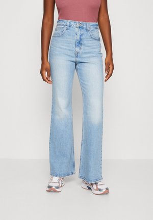 Personne portant un jean bleu clair taille haute à jambes larges, un haut sans manches mauve et des baskets blanches avec des détails roses et argentés.