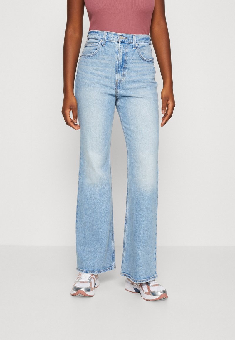 Personne portant un jean bleu clair taille haute à jambes larges, un haut sans manches mauve et des baskets blanches avec des détails roses et argentés.