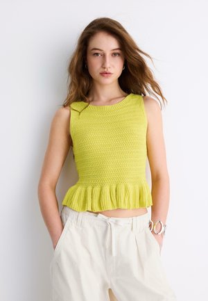 Giovane donna con capelli castano chiaro che indossa un top giallo senza maniche in maglia con orlo arricciato e pantaloni bianchi larghi con le mani nelle tasche.