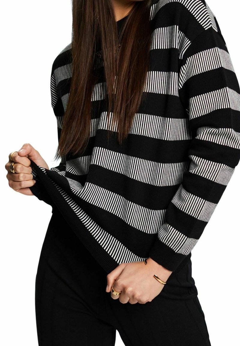 Personne aux longs cheveux bruns portant un pull rayé noir et blanc et un pantalon noir, tirant sur l'ourlet du pull, montrant des bagues en or et un bracelet.