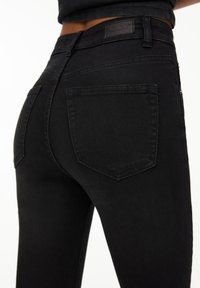 Vaqueros de mezclilla negros de talle alto con un corte ajustado, que cuentan con un diseño de bolsillo trasero y un parche de cuero con el logo en la cintura. Textura suave.