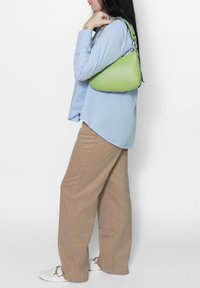 Groene leren schoudertas met een gebogen vorm, voorzien van zilveren hardware en een gladde textuur, gedragen op de schouder tegen een lichtblauw overhemd.