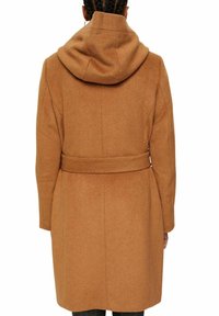 Persoon in een knie-lange camelbruine jas met een capuchon en tailleband, gezien vanaf de achterkant tegen een witte achtergrond.