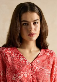 Rotes Blusenoberteil mit floralem Druck, V-Ausschnitt, Knopfdetails und kurzen Ärmeln. Der Stoff hat eine glatte Textur mit weißen Blumenmustern.