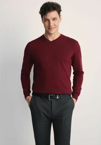 Burgundowy sweter w serek wykonany z miękkiej dzianiny, zestawiony z ciemnoszarymi spodniami i czarnym paskiem. Prosty design z długimi rękawami.