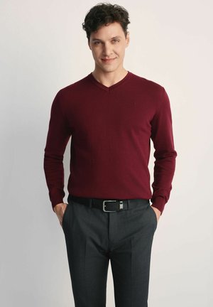 Burgundowy sweter w serek wykonany z miękkiej dzianiny, zestawiony z ciemnoszarymi spodniami i czarnym paskiem. Prosty design z długimi rękawami.