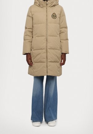 Manteau matelassé beige mi-long jusqu'aux genoux avec capuche et blason brodé, porté avec un jean bleu à jambes larges et des baskets blanches.