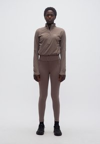 Άτομο που στέκεται, φοράει μπλούζα με μακριά μανίκια με φερμουάρ σε χρώμα τάουπε και ταιριαστά κολάν, μαζί με μαύρα αθλητικά παπούτσια, μπροστά σε απλό ανοιχτό φόντο.