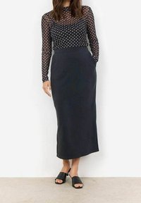 Sort lang nederdel med en glat tekstur, parret med en semi-gennemsigtig sort top med polka-prikker. Modellen bærer sorte open-toe mules.